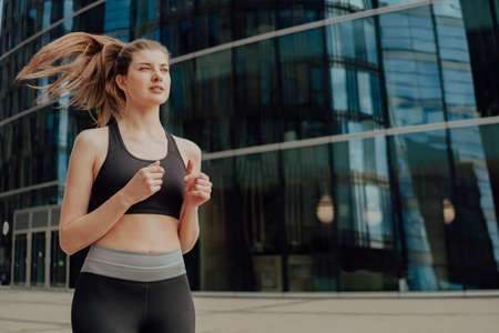 A young blonde sports girl in a black suit runs along modern housesの写真素材