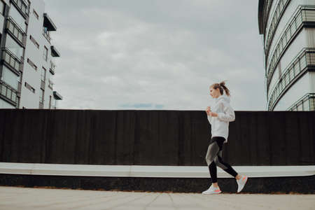 A young blonde sports girl in a black suit runs along modern houses