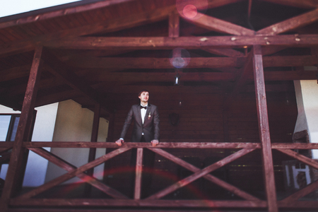 Portrait of stylish groom in a suit and bow-tie on outdoor wooden terrace of manorの写真素材