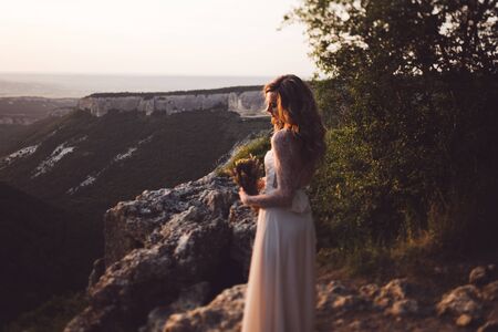 Portrait of young curly woman on a background of mountains at sunset. Wedding bouquet in handsの写真素材