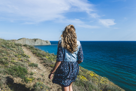 Woman in blue dress and jeans jacket walking by the sea in warm sunny weather in spring. Clear skyの写真素材