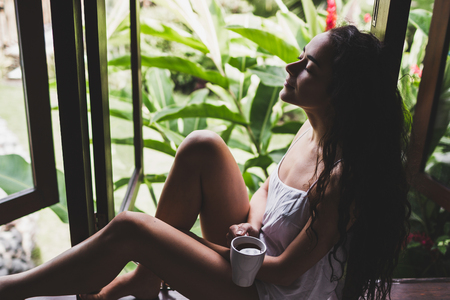 Young beautiful girl drinks morning tea on a windowsill at home in tropical gardenの写真素材