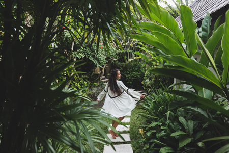 Woman walking in tropical garden in white dress. Brunette with long black hair. Fashion tunic, barefoot, enjoying natureの写真素材