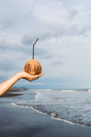 Woman's hand holding coconut at ocean coast in Bali. Blue tropical background with empty space for logo. Cold and healthy drink in summerの写真素材