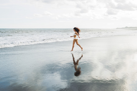 Beautiful slim tanned woman with long black hair walking alone on paradise beach in Bali at sunset. Nobody around and mirror reflection in calm water surface. Idyllic and dreamlike viewの写真素材