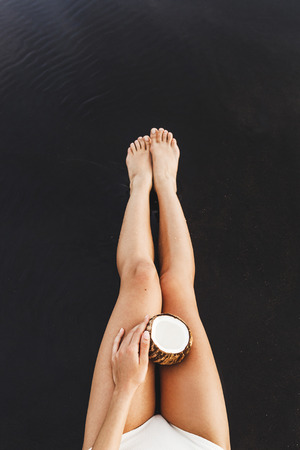 Open fresh coconut with white flesh on tanned woman legs ob black sand beach in Bali. Contrast dark background with empty space for logo and text. Island lifeの写真素材