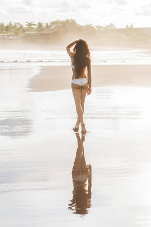 Beautiful slim tanned woman with long black hair walking alone on paradise beach in Bali at sunset. Nobody around and mirror reflection in calm water surface. Idyllic and dreamlike viewの写真素材