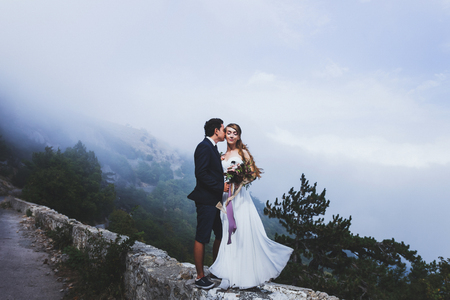Bride and groom in mountains above the clouds, posing on the edge of serpentine roadの写真素材