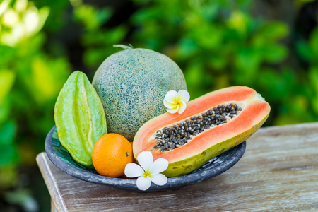 Fruit plate with fresh Bali fruits papaya, orange, melon and star fruit on wooden vintage table in tropical garden. Frangipani flowersの写真素材