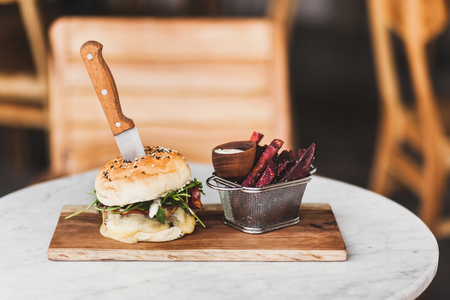 Fresh tasty burger with knife inside with sauce and fried purple sweet potato on side. On white round table on wooden boardの写真素材