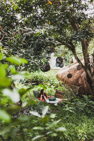 Woman in white dress relaxing in hanging hammock in bali garden. Round wooden wicker lantern hanging on treeの写真素材