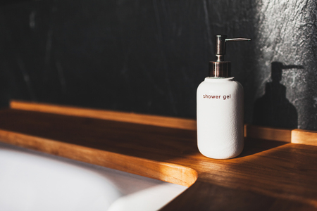 White metal bottle of shower gel on wooden edge of bath in modern hotel room with textured black wallの写真素材