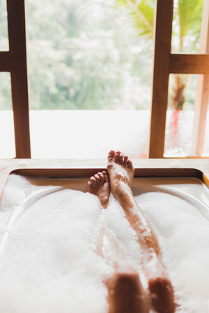 Woman legs in bath foam. Enjoying and relaxation in spa hotel room with view through windowの写真素材