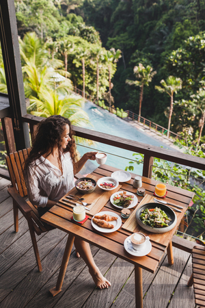 Woman enjoying breakfast on terrace with amazing jungle view. White shirt and jeans shortの写真素材
