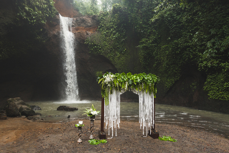 Tropical wedding ceremony with waterfall view. White arch decorated with green jungle leaves monstera and fernの写真素材