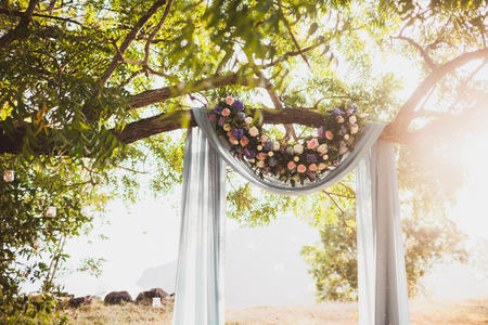 Sunset wedding ceremony, arch decorated with grey cloth hanging on big tree and rose flowers arrangementの写真素材