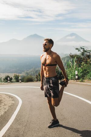Runner stretching legs and warming-up before running on the road. Summer weather, topless body and sunglasses. Using heart rate monitor.の写真素材