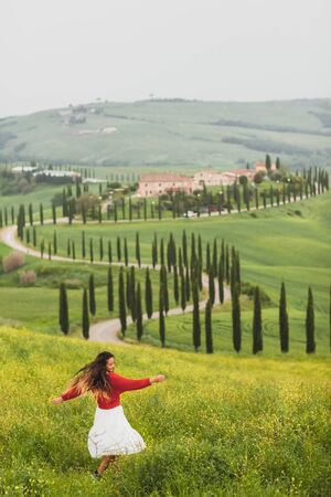 Young happy woman in red sweater and white skirt on background on spring Tuscany landscape. Dancing and enjoying view of cypress alley and yellow flowers fieldの写真素材