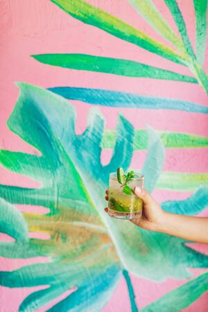 Woman is holding glass of classic mojito cocktail over trendy tropical background. Party concept.