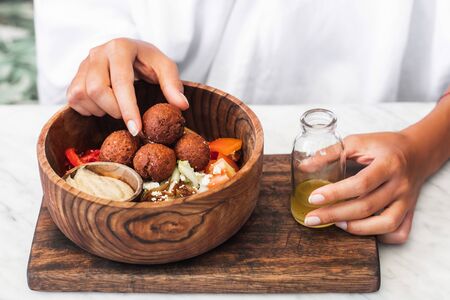 Wooden bowl with falafel balls, hummus and fresh salad. Woman hands holding olive oil on side. White...