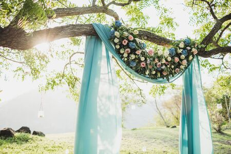 Sunset wedding ceremony arch with flower decoration and blue cloth canopy hanging on big tree. Blue hydrangea, pink roses, lily. Pastel colored.の写真素材