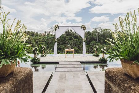 White Wedding ceremony arch in tropical jungle on edge of blue infinity swimming pool. Beautiful arch and decorations on the cliff.の写真素材