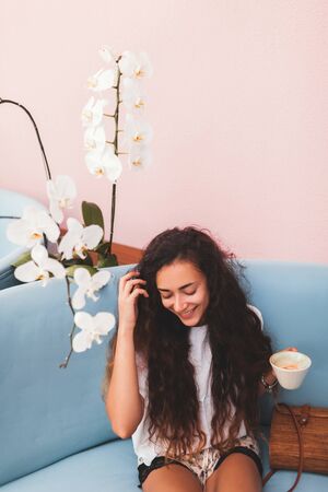 Young happy woman smiling and drinking coffee in trendy hipster cafe. Blue sofa, light coral walls,...
