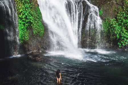 Young slim brunette woman with curly hair enjoying in lagoon of huge tropical waterfall Banyumala in Bali. Wearing in black swimsuit. Happy vacations in Indonesia. Wanderlust travel concept.の写真素材