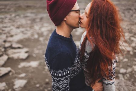 Close-up lovely portrait of young hipster couple kissing and hugging together. Traditional handmade Iceland wool sweaters, red hair and hat. Dramatic nordic stone landscape, cold weather.の写真素材