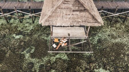 Slim tanned woman in white bikini chilling in over-reef hammock with amazing view of coral reef in...