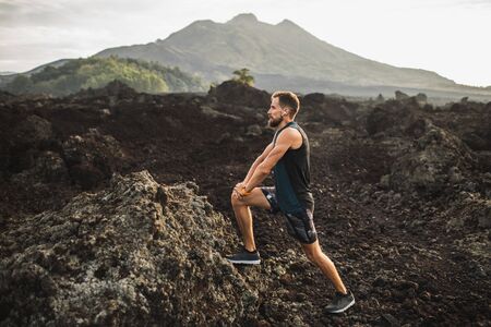 Young hipster runner with beard stretching and warming-up for trail running outdoors. Listening music in air pods. Mountain view on background.の写真素材