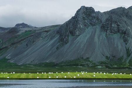 White plastic silage wrapped bales with hay on green grass hill near lake in Iceland. Farming in Scandinavia, Europe. Sunny summer weather, nature background with high rocky mountains.の写真素材