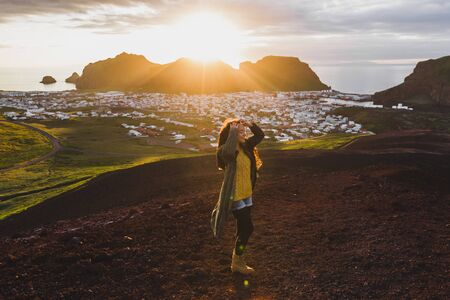 Young happy woman enjoying sunset on peak of Eldfell volcano with panoramic view of Vestmannaeyjar island in Iceland. Wanderlust travel concept. Amazing colorful sunset.の写真素材