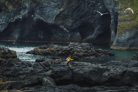 Woman walking by black stone rocky volcanic coastline of Vestmannaeyjar island in Iceland. Northern nature landscape with black beach and water caves in bay.の写真素材