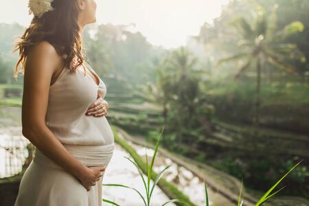 Young pregnant woman in white dress with view of Bali rice terraces in morning sunlight. View from behind. Harmony with nature. Pregnancy concept.の写真素材