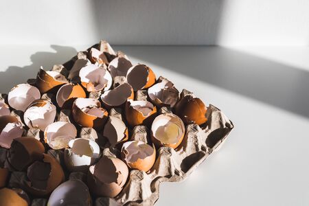 Heap of broken empty eggshell in carton box on white table. Cooking on easter concept. Unusual photo...