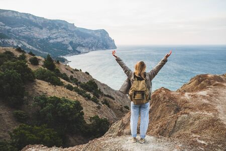 Backpacker hippie woman happy to enjoy amazing sea and mountain view from hill. View from behind. Travel concept. Unrecognizable person in jeans and beige backpack.の写真素材