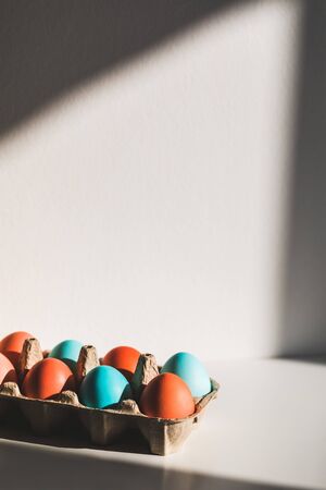 Carton box with painted blue and red chicken eggs for easter. White table and beautiful sun light....