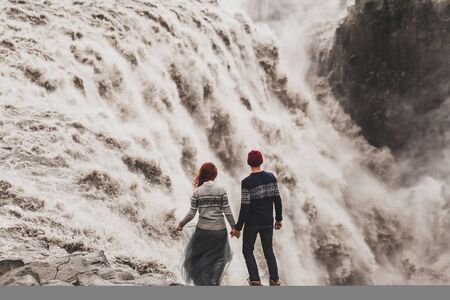 Young couple in love looking at famous Icelandic landmark Dettifoss waterfall. Traditional wool sweater, red hair, hat, gray skirt. Dramatic nordic landscape, cold weather in Iceland. View from behindの写真素材
