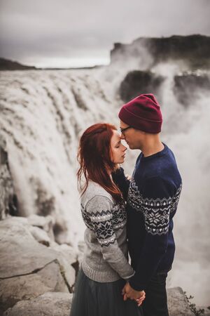 Young stylish couple in love kissing together near famous Icelandic landscape Dettifoss waterfall. Traditional wool sweaters, hat, red hair, gray skirt. Dramatic landscape, cold weather in Iceland.の写真素材