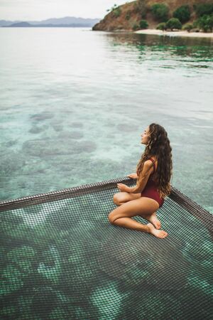 Beautiful tanned woman in red swimsuit relaxing in over-reef hammock with amazing view of coral reef...