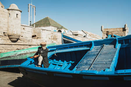 ESSAOUIRA, MOROCCO - SEPTEMBER 10, 2019: Old senior fisherman on blue wooden boat in Essaouira harbor. Stylish look, hat, beard, sunglasses and jacket. Moroccan people.のeditorial素材