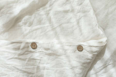 Trendy organic natural linen bedclothes with wooden buttons closeup. Bedding, morning light, bedroom style and design. Rough textile background with wrinkle.の写真素材