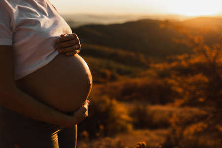 Woman hands hold belly in pregnancy outdoors closeup. Amazing warm sunset light. Maternity and childbirth concept.の写真素材