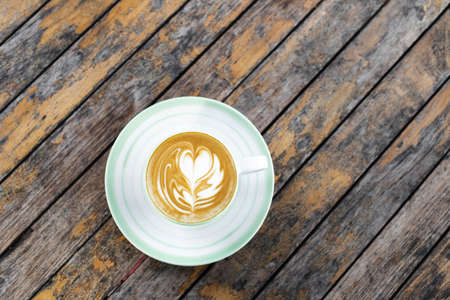Cup of fresh creamy cappuccino with latte art on foam. Background of wooden table with shabby aged...