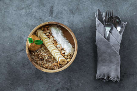Wooden bowl with healthy smoothie bowl and cutlery on gray concrete background. Banana, peanut butter, coconut flakes and nuts ingredients.の写真素材