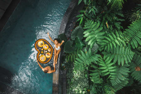 Travel happy couple in love eating floating breakfast in jungle swimming pool. Awakening in morning. Black rattan tray in heart shape, Valentines day or honeymoon surprise, view from above.の写真素材