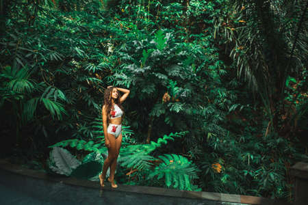 Beautiful slim brunette woman in white swimsuit on edge luxury swimming pool outdoors in Jungle. Bali.の写真素材