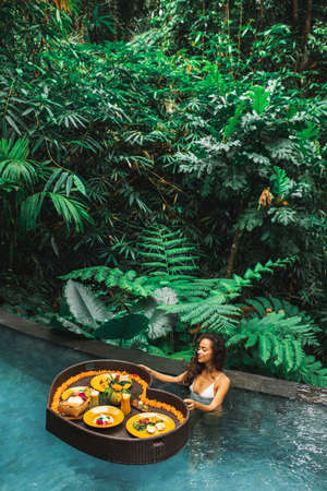 Woman relaxing and having floating breakfast in jungle pool on luxury villa in Bali. Valentines day...