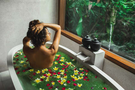Beautiful young woman enjoying in spa, view from behind. Luxury stone bath tub with jungle view in...